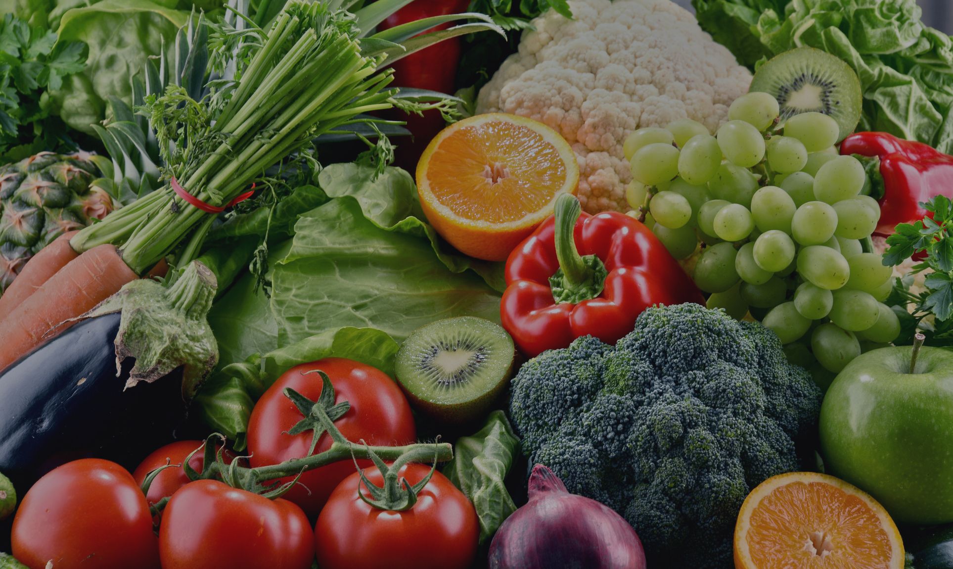 Eine lebendige Auswahl an frischem Gemüse und Obst, darunter Brokkoli, Tomaten, Paprika, Trauben und Orangen auf dunklem Hintergrund.